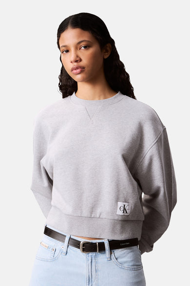 Pull-over gris à col rond de Calvin Klein, avec un logo de marque et associé à une ceinture sur un jean.