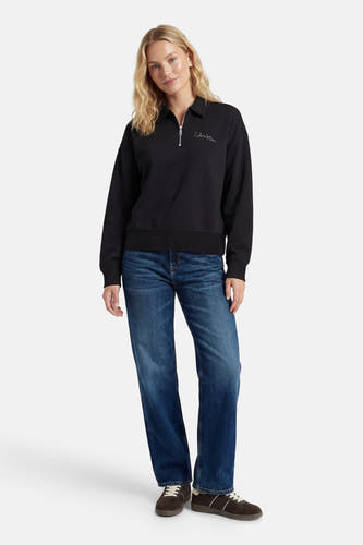 Pull-over noir de Calvin Klein avec une fermeture éclair et le logo sur la poitrine, associé à des jeans bleus et des chaussures marrons.