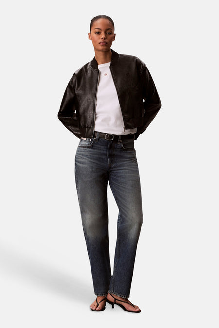 T-shirt blanc Calvin Klein, à manches courtes, porté sous un blouson bomber en cuir noir, associé à un jean bleu et des sandales noires.