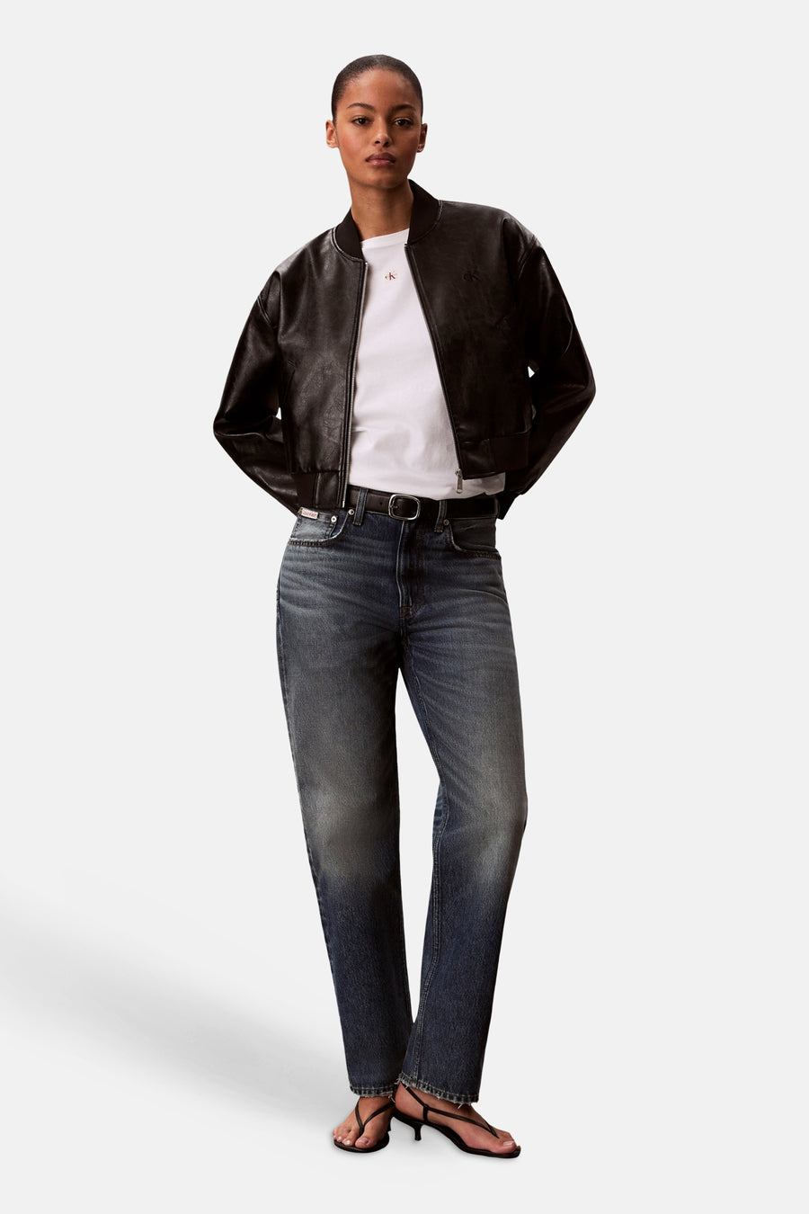 Wit T-shirt van Calvin Klein, met korte mouwen, gedragen onder een zwart leren bomberjack, gecombineerd met blauwe jeans en zwarte sandalen.
