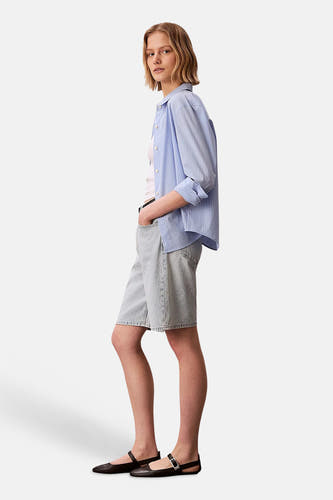 Lichtblauwe denim short van Calvin Klein, gedragen met een gestreept overhemd en zwarte ballerina's in zijaanzicht.
