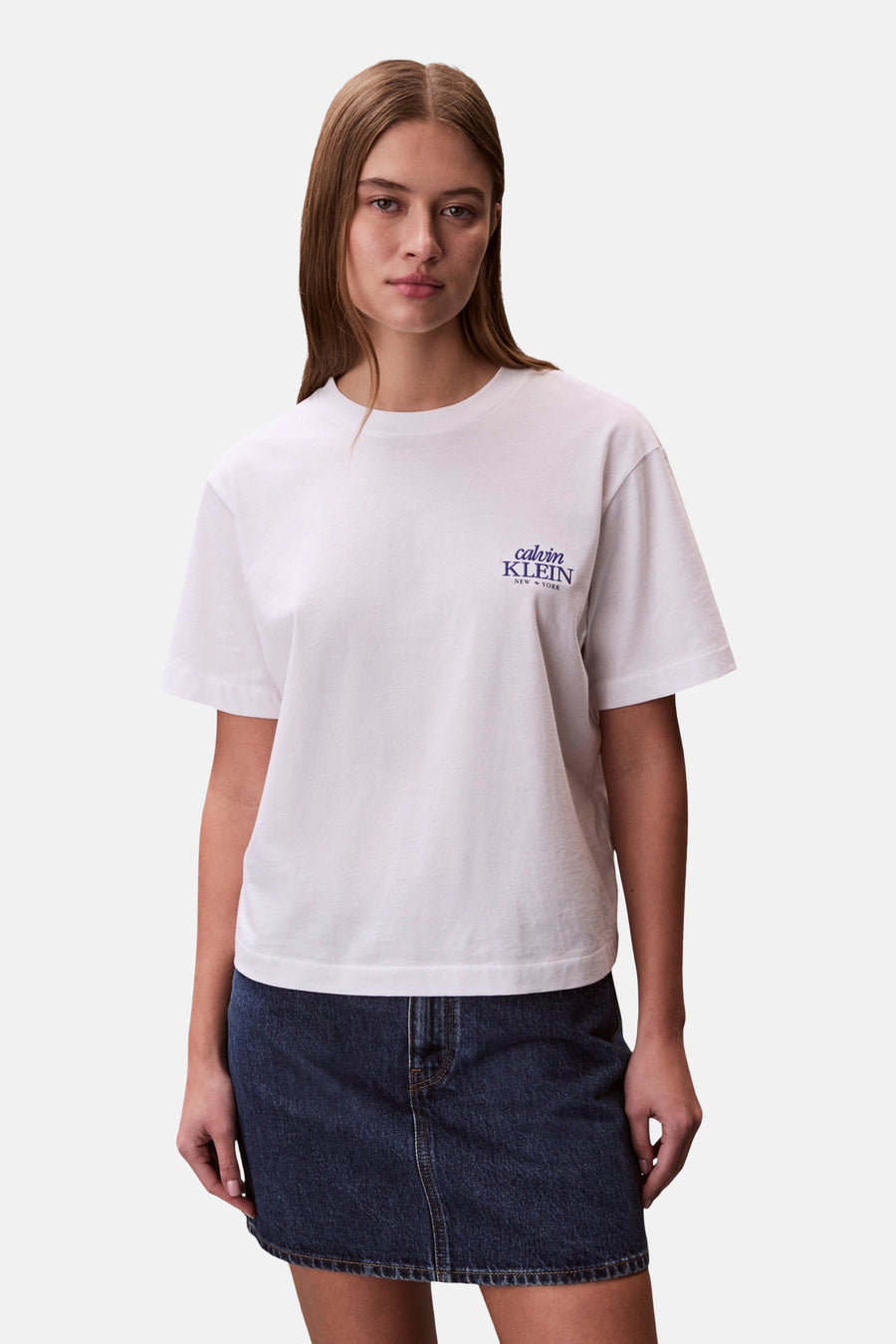 T-shirt blanc à manches courtes Calvin Klein, avec un petit logo violet sur la poitrine et porté avec une jupe en jean bleue.