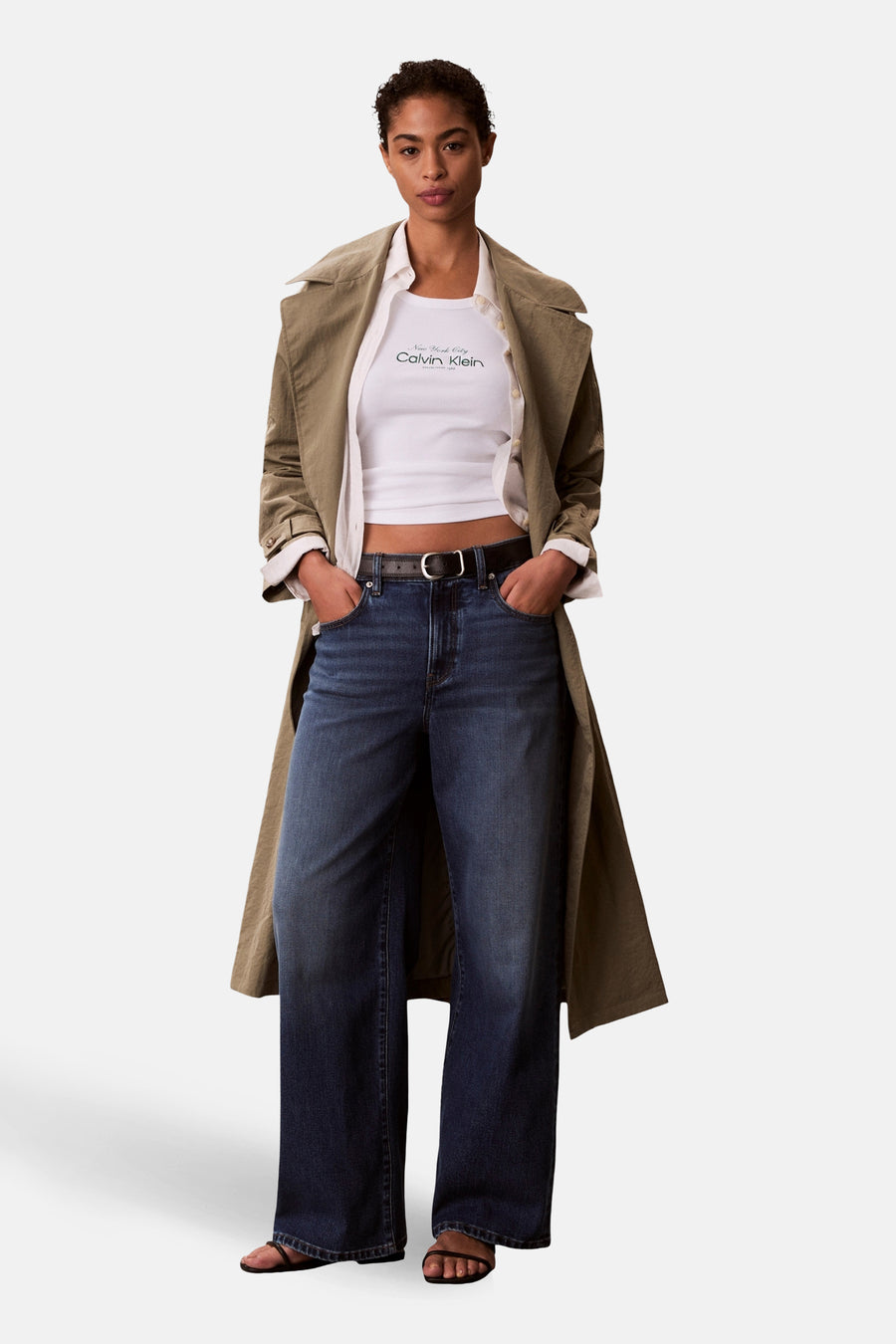 Witte tanktop van Calvin Klein, met een ronde hals en logo-opdruk, gecombineerd met een kaki trenchcoat, wit overhemd en donkere jeans.

