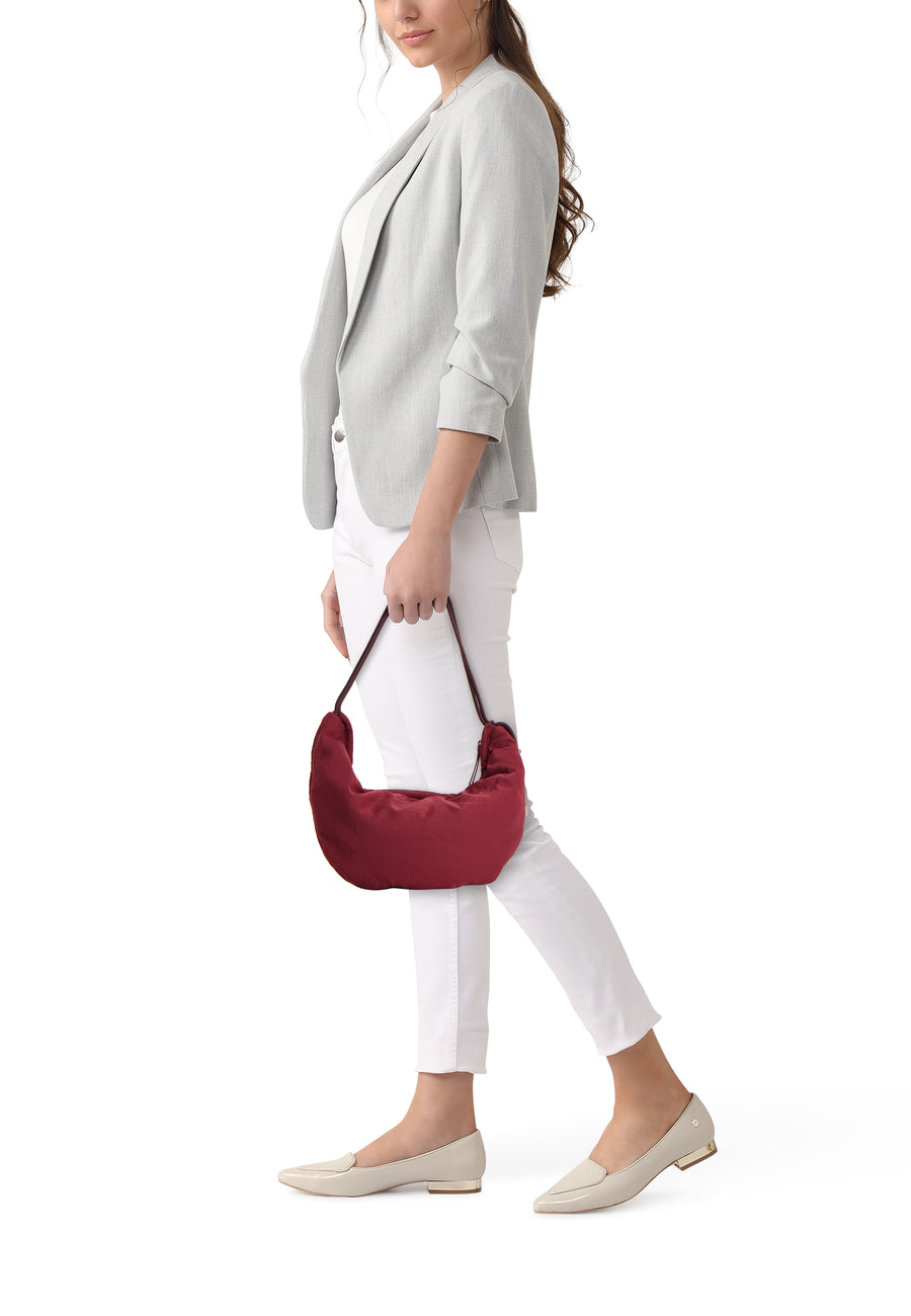 Bordeaux schoudertas van Calvin Klein, gedragen door een vrouw met een grijze blazer, witte broek en beige loafers.
