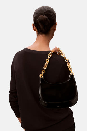 Sac à dos noir de Calvin Klein avec une chaîne dorée comme fermeture contre un fond uni.