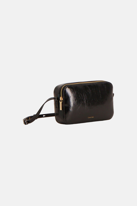 Zwarte glanzende schoudertas van Calvin Klein met goudkleurige rits, verstelbare riem en klein logo, schuin gepresenteerd.