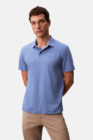 Man in blauw poloshirt met kaki broek, handen achter de rug, op witte achtergrond.