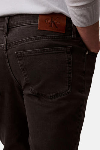 Zwarte slim fit jeans van Calvin Klein, met bruin leren merklabel, achterzak en een hand in de zak.

