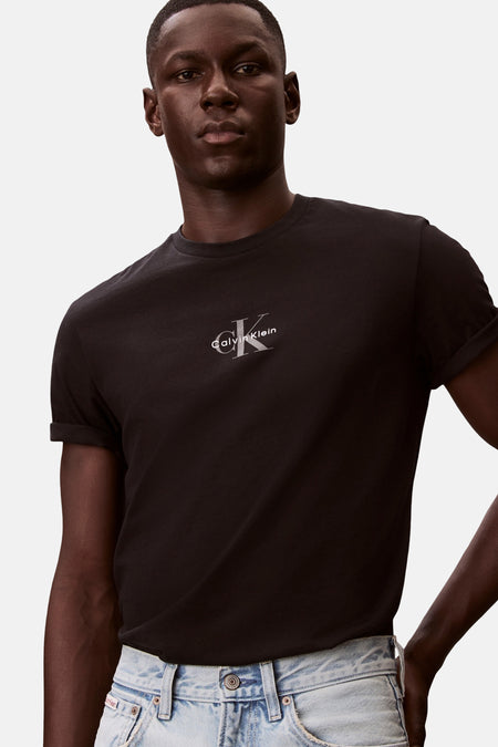 Zwart T-shirt met korte mouwen van Calvin Klein, met zilverkleurig logo op de borst en opgerolde mouwen, gedragen door een man met lichte jeans.