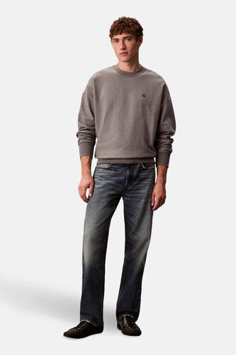 Grijze sweater van Calvin Klein, met een kleine CK logo op de borst, gecombineerd met donkerblauwe jeans en zwarte schoenen.
