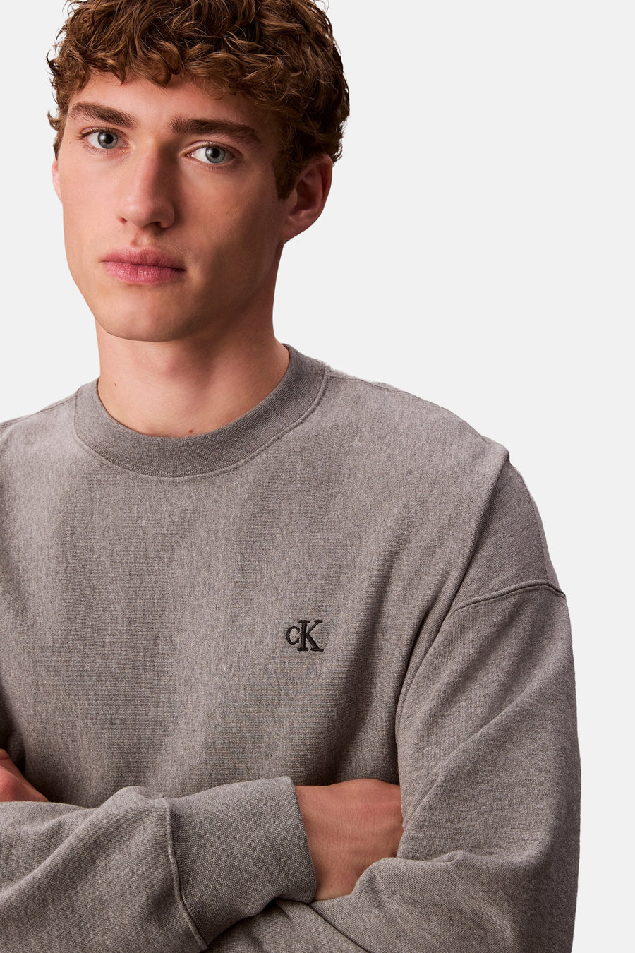Een man met krullend haar in een grijze Calvin Klein trui met gekruiste armen.