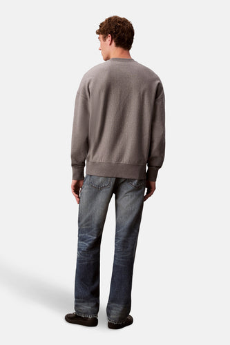 Man in bruine sweater en blauwe jeans, staand op witte achtergrond.