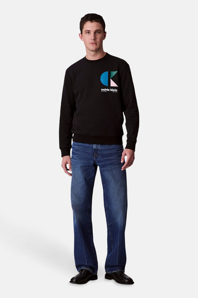 Zwarte sweater van Calvin Klein met een kleurrijk logo op de borst, gecombineerd met blauwe jeans en zwarte schoenen.
