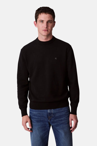 Pull-over noir de Calvin Klein avec un col rond, porté avec un jean bleu.
