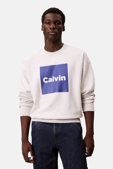 Grijze sweater van Calvin Klein, met een blauw vierkant met de tekst "Calvin" erop, gecombineerd met donkerblauwe jeans.
