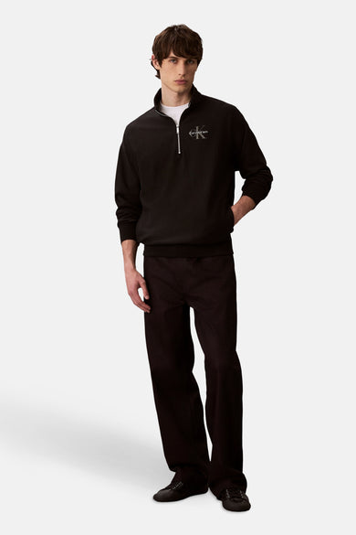 Pull-over noir de Calvin Klein avec fermeture éclair et logo sur la poitrine, associé à des pantalons et des chaussures noirs.