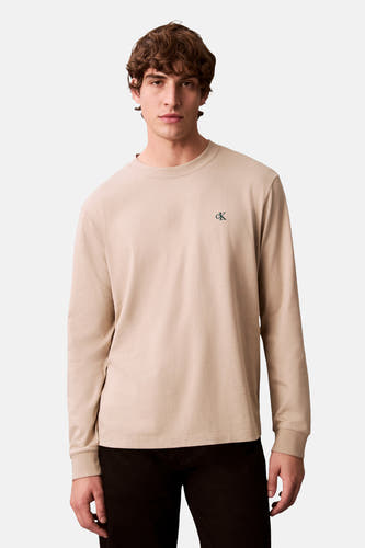 Beige T-shirt met lange mouwen van Calvin Klein, met klein logo op de borst, gedragen met zwarte broek.
