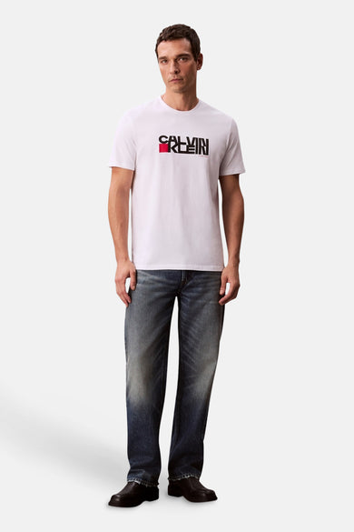 Wit T-shirt met korte mouwen van Calvin Klein, met zwart en rood logo op de borst, gecombineerd met jeans en zwarte schoenen.
