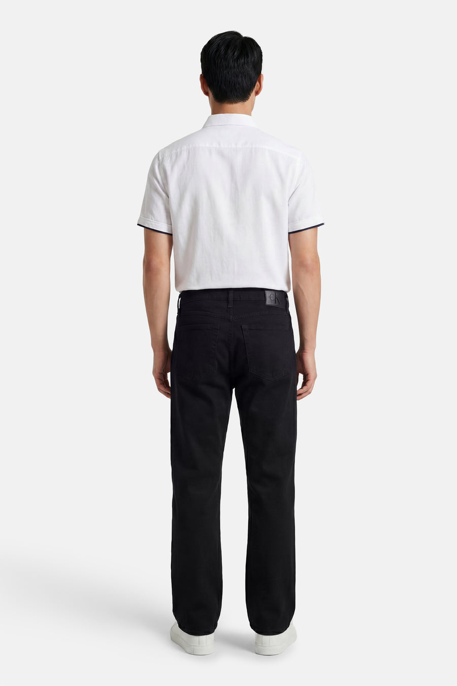 Zwarte straight fit spijkerbroek van Calvin Klein, met zichtbare CK-patch, gedragen door een man met een wit shirt en sneakers, achterkant.