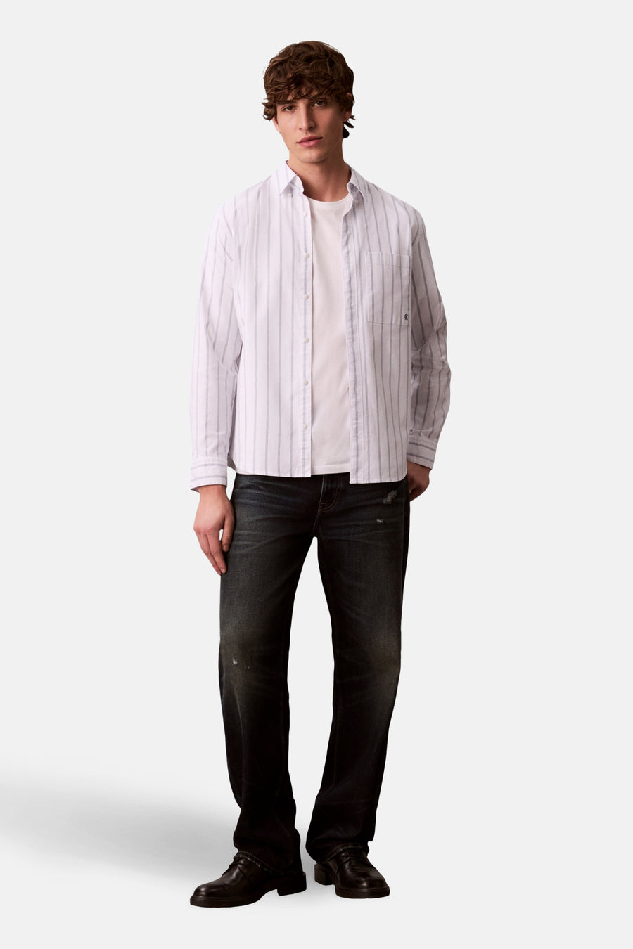 Wit overhemd met lange mouwen van Calvin Klein, gedragen over een wit T-shirt en gecombineerd met donkere jeans en zwarte schoenen.