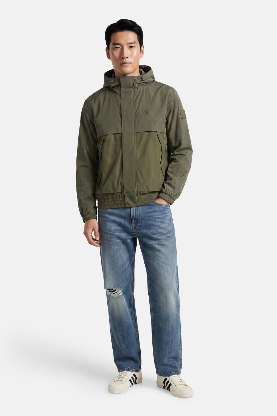 Kaki korte jas van Calvin Klein met capuchon, rits, borstzakken en logo op borst, gedragen met blauwe jeans en witte sneakers, front view.