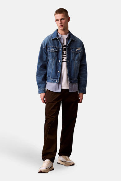 Chemise bleue à manches longues de Calvin Klein, portée sous une veste en denim, avec un pantalon marron et des baskets crème.