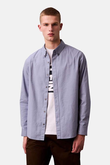 Blauw overhemd met lange mouwen van Calvin Klein, gedragen over een wit T-shirt en bruine broek.
