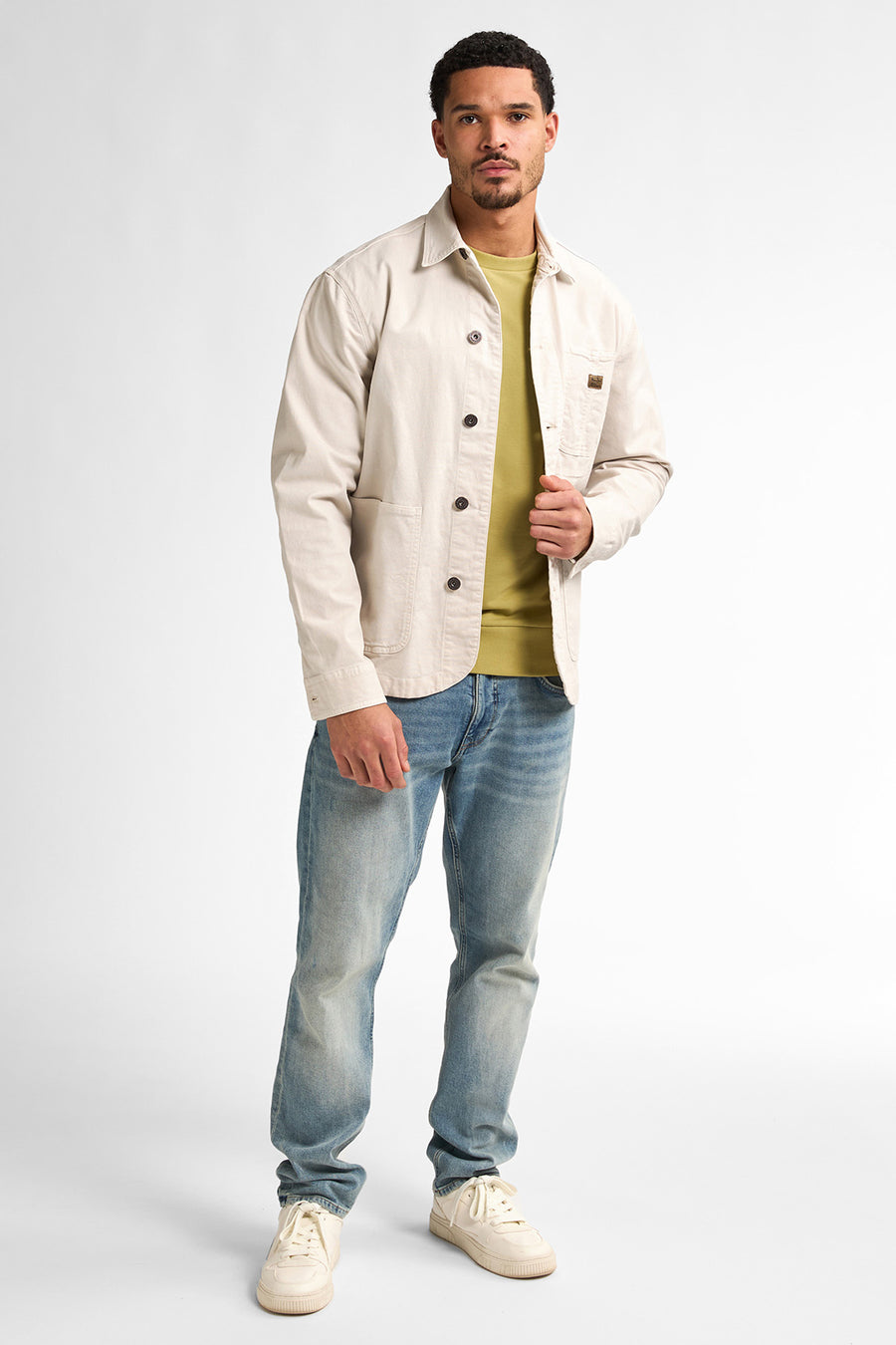 Man met beige jack, groene trui, jeans en sneakers, staand tegen een witte achtergrond.