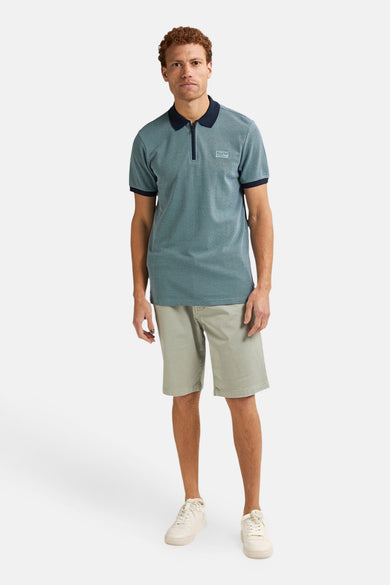 Blauwe polo met korte mouwen van Petrol Industries, met korte rits en gedragen met kaki shorts en sneakers.

