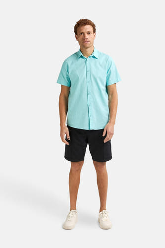 Turquoise hemd met korte mouwen van Petrol Industries, gedragen met zwarte shorts en crèmekleurige sneakers.
