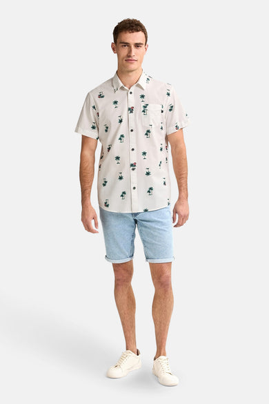 Chemise blanche à manches courtes de Petrol Industries, avec des palmiers et portée avec des shorts en denim et des baskets blanches.