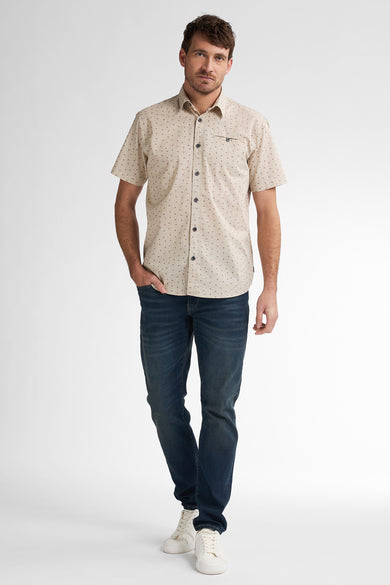 Chemise beige à manches courtes de Petrol Industries, avec un petit motif à carreaux, portée avec des jeans foncés et des baskets blanches.