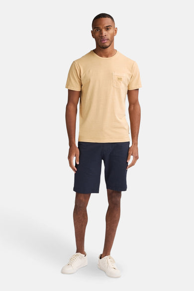 T-shirt jaune à manches courtes de Petrol Industries, avec une poche po poitrine et porté avec des shorts bleus foncés et des baskets blanches.