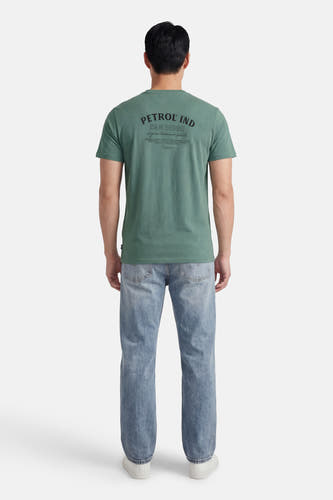Groen T-shirt met korte mouwen van Petrol Industries, met bedrukte tekst op de achterkant, gecombineerd met blauwe jeans en witte schoenen.