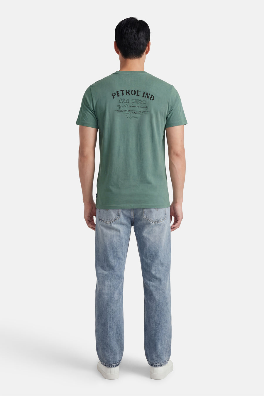 Groen T-shirt met korte mouwen van Petrol Industries, met bedrukte tekst op de achterkant, gecombineerd met blauwe jeans en witte schoenen.