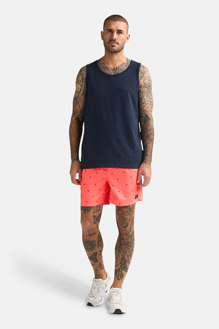 Débardeur bleu de Petrol Industries, porté par un homme tatoué avec un short rouge corail à motif d'ancre et des baskets blanches.