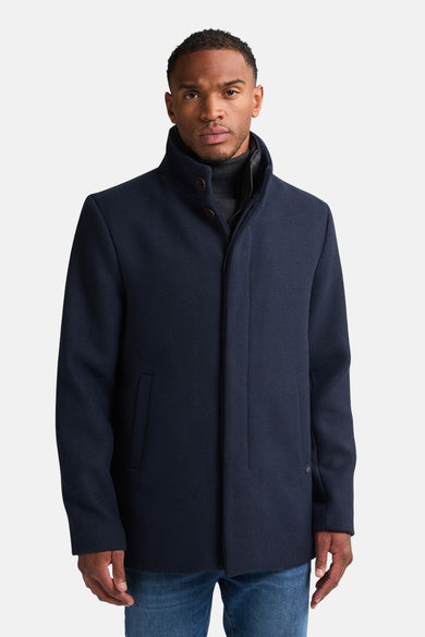 Blauwe lange jas van Petrol Industries, met opstaande kraag en een man met grijze coltrui en blauwe jeans