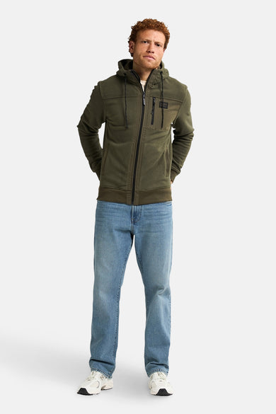 Groene sweater gilet van Petrol Industries, met rits en capuchon, gedragen met blauwe jeans en witte sneakers.
