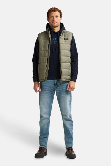 Kaki bodywarmer van Petrol Industries, gedragen over een marineblauwe hoodie en blauwe jeans, met bruine veterschoenen.