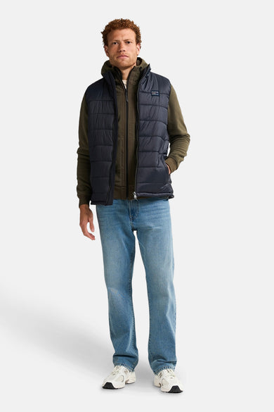 Gilet de chauffe body noir de Petrol Industries, porté sur un hoodie vert et une veste marron, avec des jeans bleus et des baskets blanches.