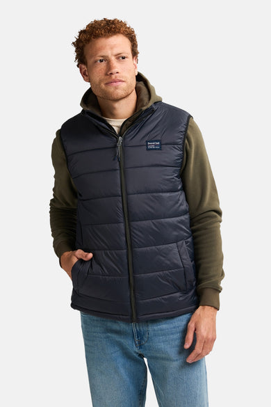 Gilet chauffant noir de Petrol Industries avec des coutures verticales, porté sur un hoodie vert et un pantalon en denim.