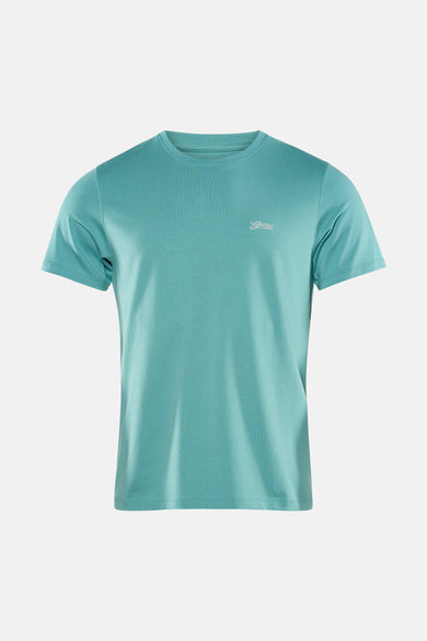 T-shirt vert à manches courtes Guess®, avec petit imprimé texte sur la poitrine, combiné avec un jean foncé et des baskets beiges.