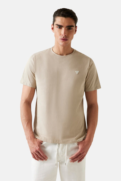 T-shirt beige à manches courtes de Guess, avec un petit logo sur la poitrine, porté avec un pantalon blanc.
