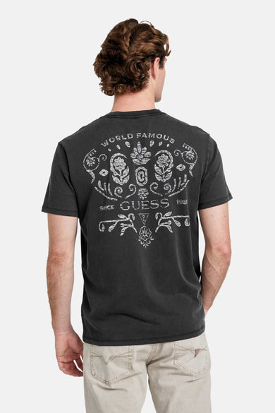 T-shirt gris à manches courtes de Guess®, avec logo sur la poitrine, associé à un pantalon léger et des baskets blanches.