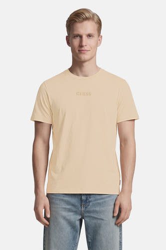 Beige T-shirt van Guess met ton-sur-ton GUESS-logo op de borst, gedragen door een man met lichtblauwe jeans.