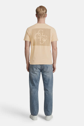 Beige T-shirt van Guess met ton-sur-ton logo op de rug, gedragen door een model met lichte jeans met studs en witte sneakers, achterkant.