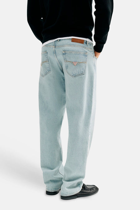 Rechte lichtblauwe denim jeans van Guess, met bruine leren patch en driehoekig logo op de rechter achterzak, gedragen met zwarte instappers, rugaanzicht.