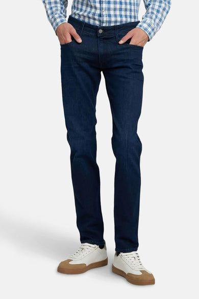Jean slim fit bleu marine de REPLAY, avec des mains dans les poches, associé à une chemise à carreaux et des baskets à semelles brunes.