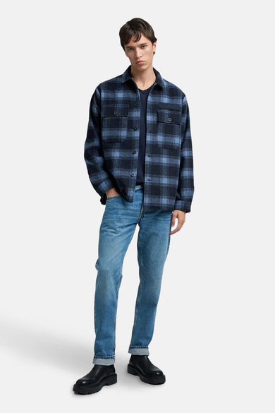 Blauwe rechte jeans van REPLAY, gedragen met een donkerblauw shirt en een blauw-zwart geruit overhemd.