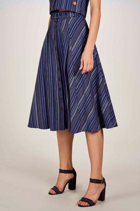 Jupe 3/4 bleue avec des rayures blanches et dorées de Le Fabuleux Marcel de Bruxelles, avec des boutons à l'avant, portée avec un haut rayé et des talons noirs.